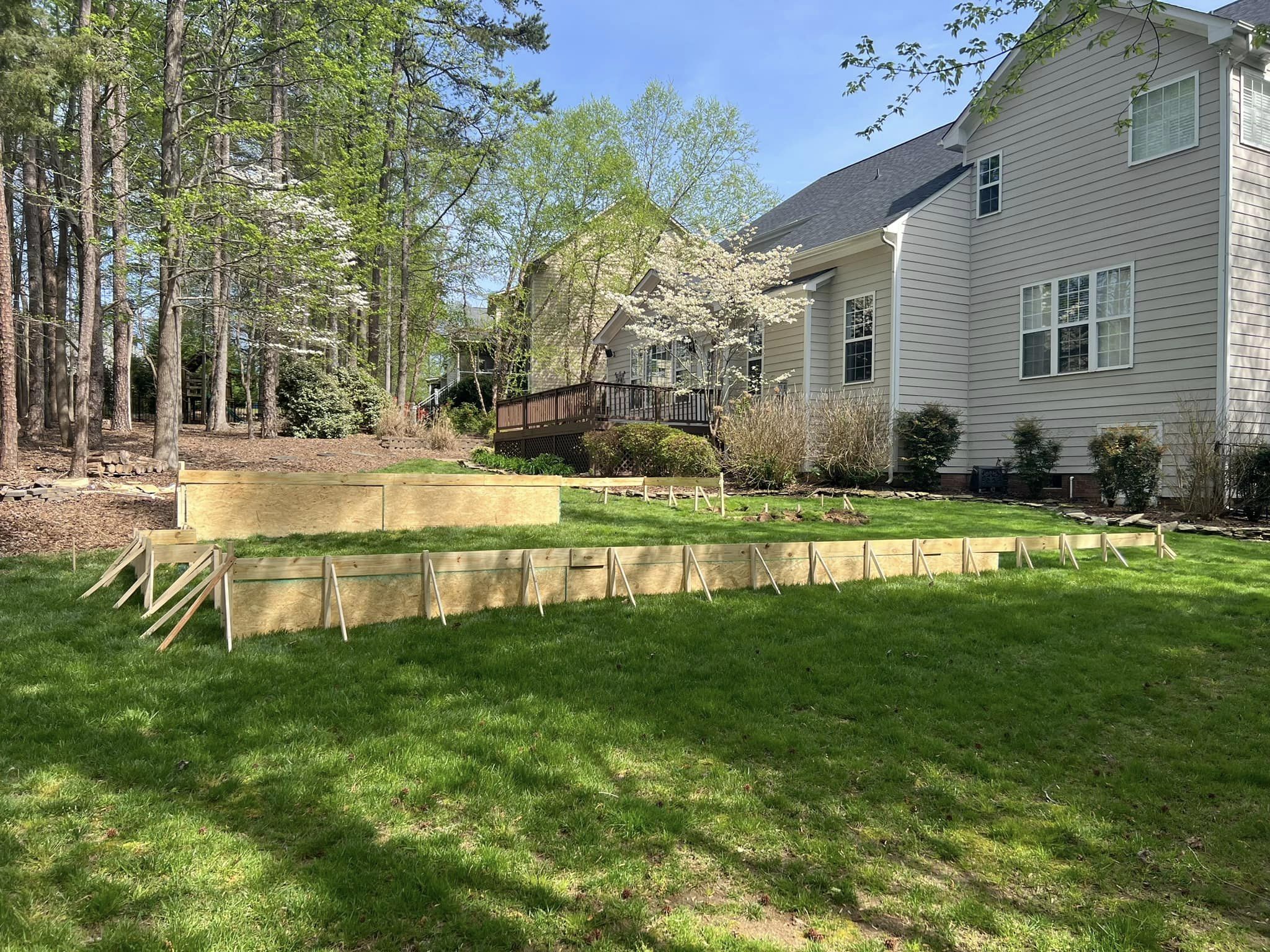 A backyard with a wooden frame structure set up on a grassy area. The house is in the background, surrounded by trees and spring foliage. The sky is clear and sunny.