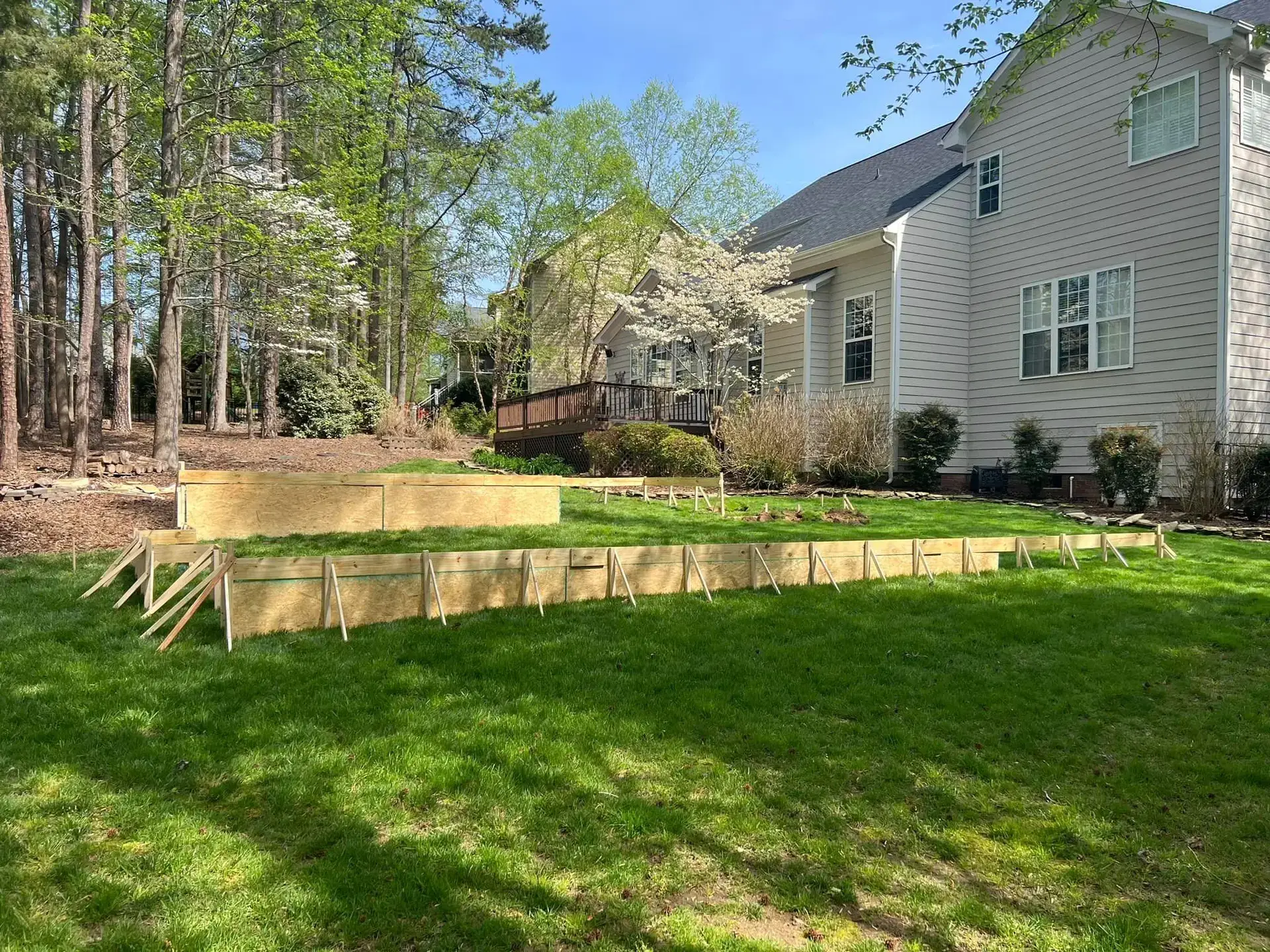 A backyard with a wooden frame structure set up on a grassy area. The house is in the background, surrounded by trees and spring foliage. The sky is clear and sunny.