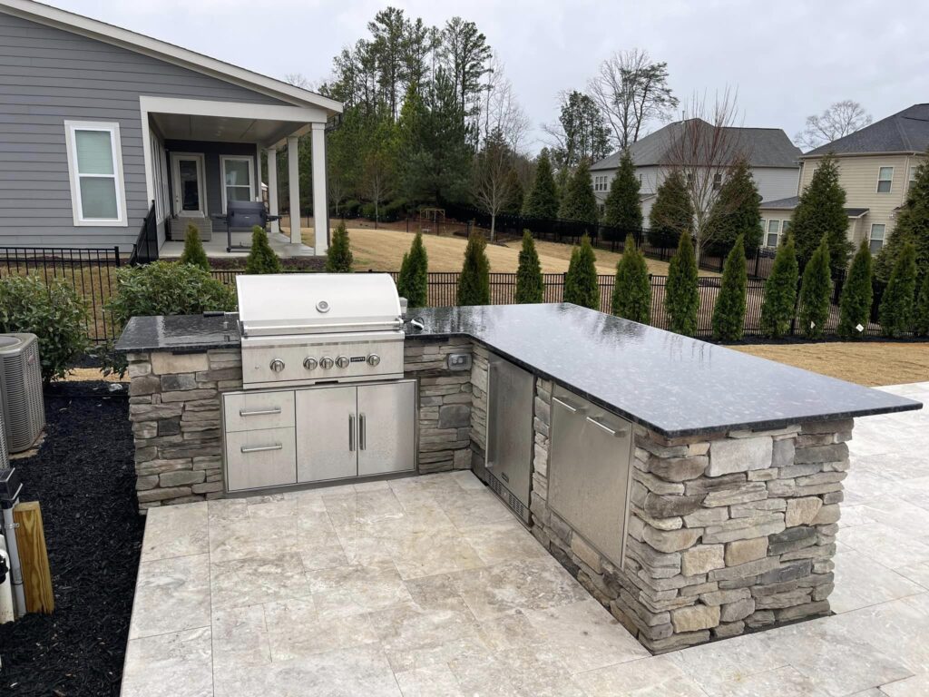 Outdoor kitchen with a built-in grill and stone counter in a backyard. The counter has stainless steel appliances and is set on a stone base. Nearby are a lawn and trees, with a modern house and a patio in the background.