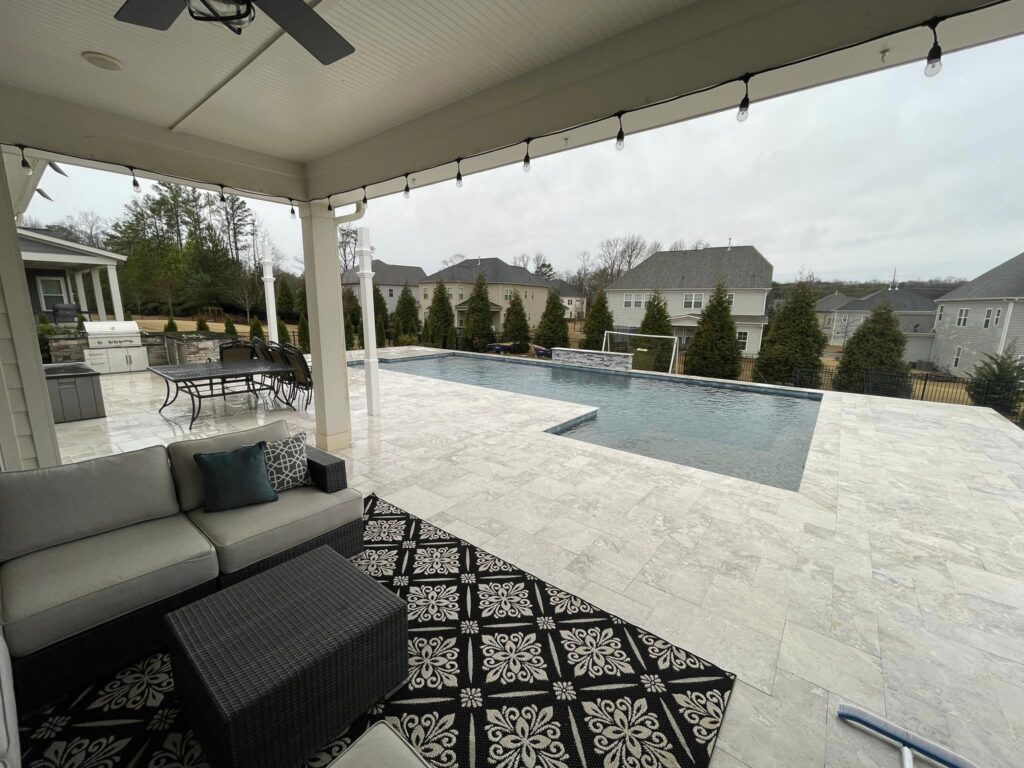 View of a luxurious backyard with a marble-tiled patio, a dark-bordered rectangular pool, a cozy seating area with a patterned rug, and outdoor dining space. String lights hang overhead, and the area is surrounded by trees and residential houses.