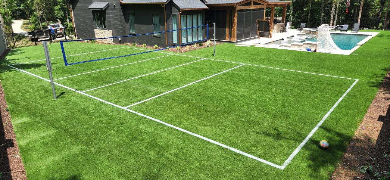 A backyard features an artificial turf area with a setup for volleyball and a marked court. In the background, there is a modern house, a patio area with seating, and an in-ground swimming pool surrounded by trees.