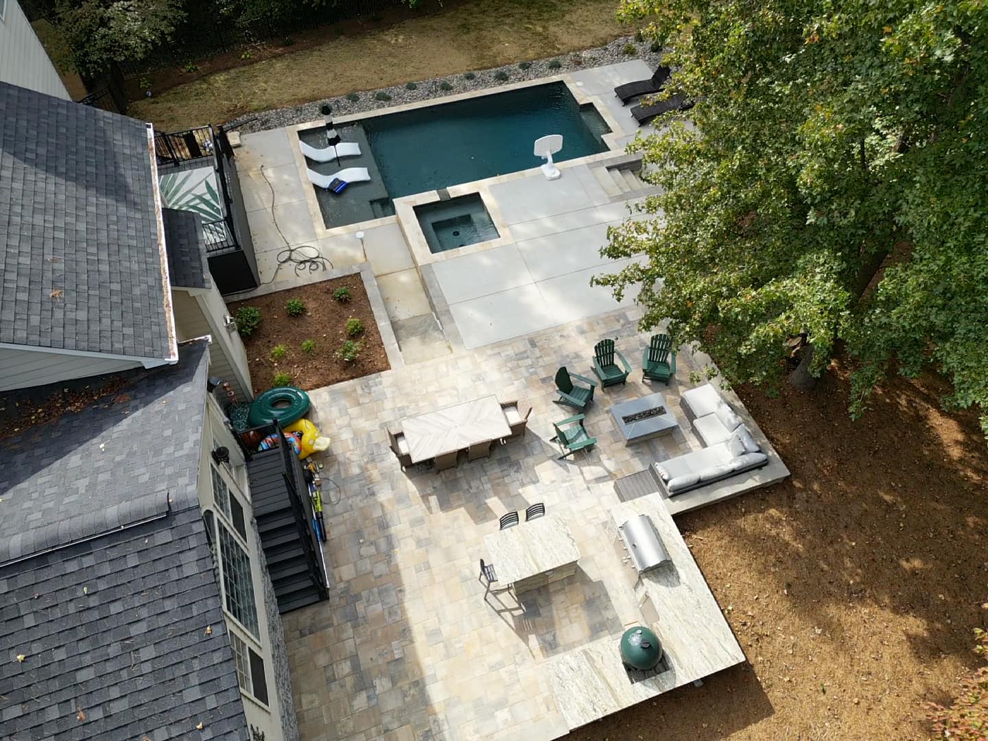 Aerial view of a backyard featuring a rectangular pool and hot tub. Surrounding the pool are lounge chairs. Adjacent is a patio with a dining table, chairs, and a grilling area. Trees and a grassy area complete the setting.