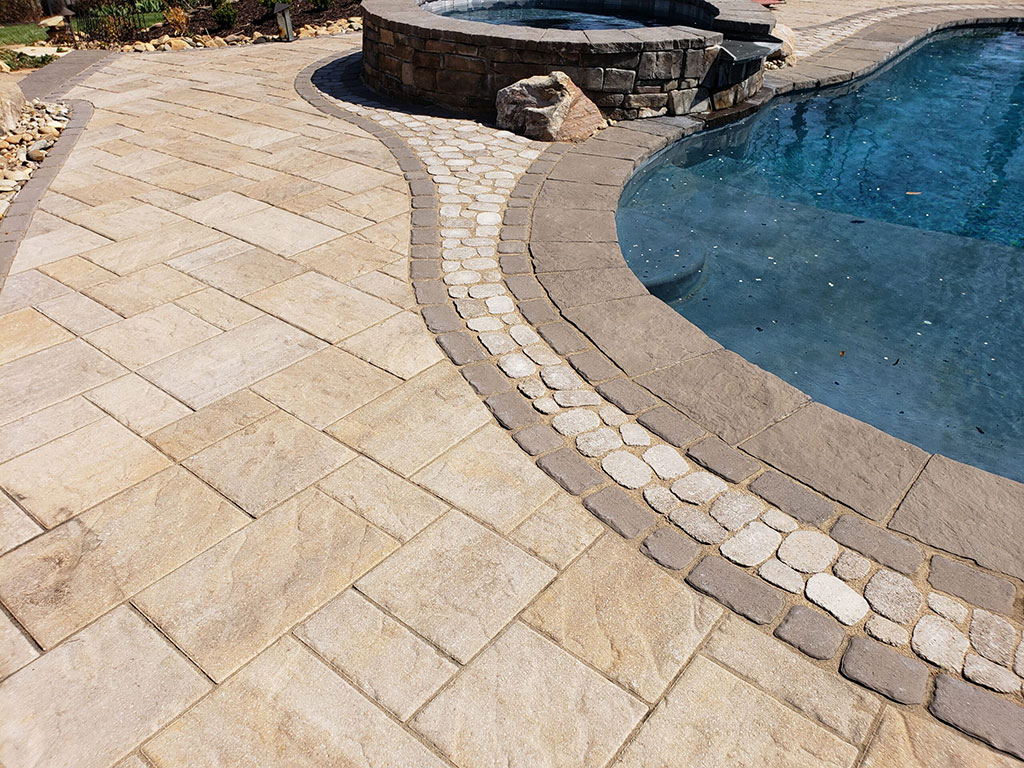 A stone-paved poolside area featuring rectangular tiles and a decorative curved border of small round stones. The pool has a curving edge and clear blue water. A raised spa is visible in the background.