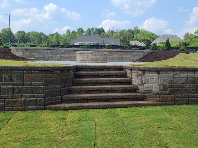 Stone steps lead up to a raised platform surrounded by neatly trimmed grass and landscaping. In the background, suburban houses are visible under a partly cloudy sky.